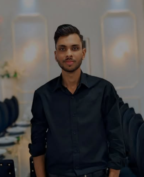 Man in a black dress shirt standing in a formal setting with banquet chairs and soft lighting.